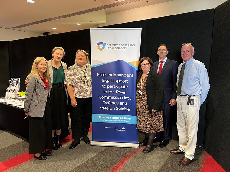 Six friendly looking people standing next to a banner for the Defence and Veterans Legal Service